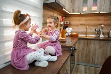 Mor kıyafetler giymiş iki genç kız mutfak tezgahında şakacı bir şekilde etkileşim halindeler. Kahkahaları sıcak boşluğu doldurur, pencereden süzülen yumuşak güneş ışığıyla aydınlanır..