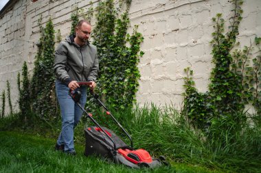 Huzurlu bir arka bahçede, bir adam elektrikli çim biçme makinesi kullanıyor, canlı yeşil çimlerle ustalıkla ilgileniyor. Uzun sarmaşık beyaz duvara tırmanıyor ve bu peyzaj işi için sakin bir zemin oluşturuyor..