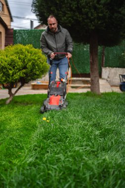 Bir adam sıcacık bir arka bahçede canlı yeşil bir çim biçiyor, sıcak güneşin ve taze kesilmiş çimlerin tadını çıkarırken aynı zamanda temiz bir açık alanı koruyor..