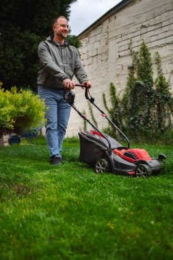 Bereketli bir bahçe ortamında, bir adam etkin bir şekilde bakımlı yeşil bir çim biçiyor. Gri bir ceket ve kot pantolon giyer, bulutlu bir öğleden sonra açık hava alanının tadını çıkarır..