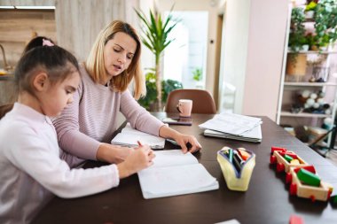 Bir anne, kızına ahşap bir masada ödevlerinde yardım eder..