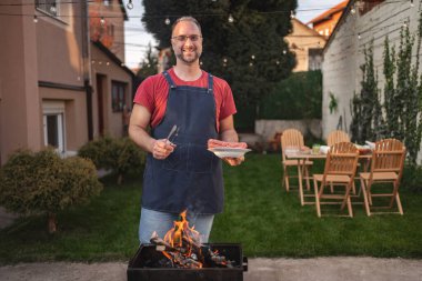 Neşeli bir adam lezzetli yemekler ve gülümsemelerle arka bahçedeki barbeküye hazırlanıyor..