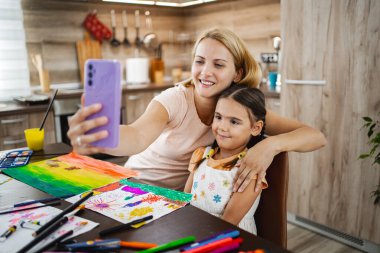 Bir anne ve kız, renkli sanat projelerinin ortasında, rahat bir mutfak ortamında selfie çekerken gülümsüyor..