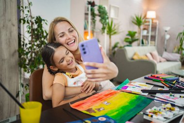 Bir anne ve kızı, canlı sanat malzemeleri ve renkli eserleriyle çevrelenmiş bir şekilde selfie çekerken mutlu bir an yaşarlar..