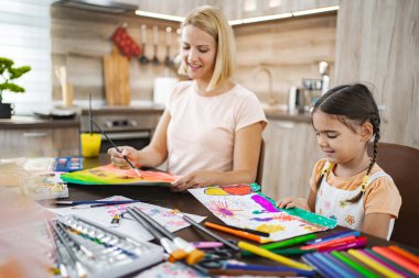 Bir anne ve kızı sanat malzemeleriyle dolu bir masada otururken canlı resimler çizerek neşeli bir sanat aktivitesine girişiyor..