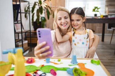 Gülümseyen bir anne ve kızı, masada canlı oyuncaklarla çevrili bir selfie çekerek birlikte eğlenceli bir anın tadını çıkarırlar..