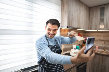 Bir adam mutfakta dikiliyor, elinde akıllı bir telefonla kendi fotoğrafını çekiyor. Mutfak, arka planda modern aletler ve çatal bıçaklarla iyi aydınlatılmış..