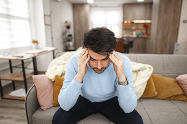 Adam oturma odasındaki kanepede oturuyor, elleriyle başını tutuyor ve büyük bir baş ağrısı hissediyor..