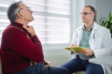 Yaşlı bir adam semptomlarını anlatırken bir doktor dikkatlice dinler..