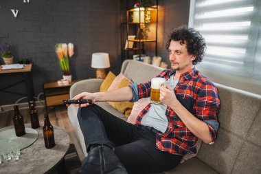 Rahatlamış genç adam evde televizyon izlerken bira içiyor.
