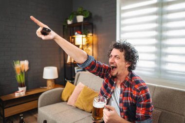 Genç yetişkin adam TV 'de maç izlemekten, bira içmekten ve kanal değiştirmekten zevk alıyor.