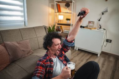 Adam evde TV 'de maç izlerken coşkuyla tezahürat yapıyor, elinde bira ve uzaktan kumandayla.