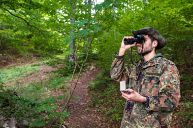 Orman bekçisi yemyeşil bir ormanda dürbün ve pusula kullanarak geziniyor.