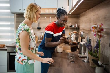 İki arkadaş şık bir mutfakta kahve hazırlarken keyifli bir anın tadını çıkarıyorlar. Modern aletler ve sıcak dekorlarla çevrili kahkahalar ve sohbetleri paylaşıyorlar..