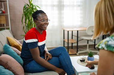 İki arkadaş sıcak bir oturma odasında keyifli bir anın tadını çıkarırlar. Biri neşeyle gülerken, diğeri etrafı bitkiler ve renkli yastıklarla çevrili kahve servisi yapıyor..