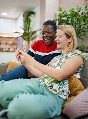 İki arkadaş, rahat bir oturma odasında mobil bir cihaz üzerinde birbirleriyle iletişim kurarken kahkahalar ve gülümsemeler paylaşırlar. Sıcak atmosfer onların bağlantısını ve eğlencesini arttırır..