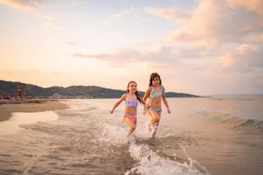 İki kız, sakin bir sahilde mutlu bir şekilde dalgaların arasında koşuyor..