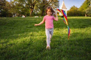 Parlak bir gülümsemesi olan genç bir kız, hafif esintide dalgalanan canlı bir gökkuşağı uçurtmasıyla yemyeşil bir parkta koşuyor. Güneş onun neşeli keşfine sıcak bir ışık tutuyor..