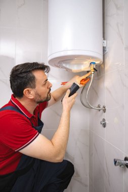 Tesisatçı banyodaki elektrikli su ısıtıcısını el feneri ve penseyle kontrol ediyor.
