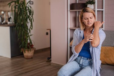 Kanepede oturan, kaşınan kolunu kaşıyan, evinde deri iltihabından muzdarip bir kadın.