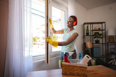 Mutlu temizlikçi kadın pencereyi açıyor ve kulaklıkla müzik dinliyor, ev temizliği hizmeti konsepti