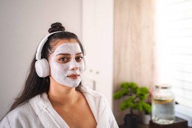 Güzellik maskesi takan ve kulaklıkla müzik dinleyen genç bir kadın evde bir anlık rahatlığın tadını çıkarıyor.