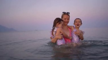 Anne ve iki kız mayo giyip, güzel bir gün batımında sakin denizde eğleniyorlar.