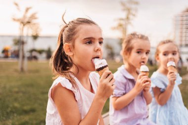 Üç küçük kız parkta dondurma yiyorlar, yaz gününün tadını çıkarıyorlar.