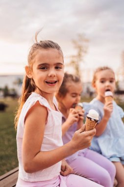 Üç küçük kız bankta oturmuş dondurma yiyorlar ve kameraya gülümsüyorlar.