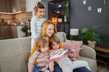 Anne ve üç kızı oturma odasında kanepede oturmuş annelerinin bir hikaye okumasını dikkatle dinliyorlar..