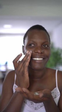 Genç Afrikalı Amerikalı kadın nemlendirici yüz kremi sürüyor, gülümsüyor ve kameraya bakıyor.