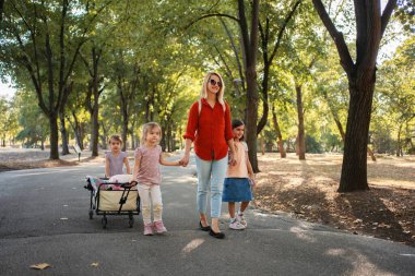 Anne ve kızları parkta yürümekten zevk alıyorlar, oyuncak ve battaniyelerle yük arabalarını çekiyorlar.