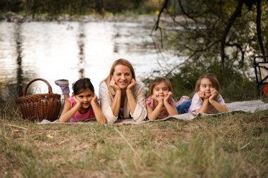 Anne ve üç kızı nehir kenarındaki battaniyeye uzanmış piknik yapıyorlar.