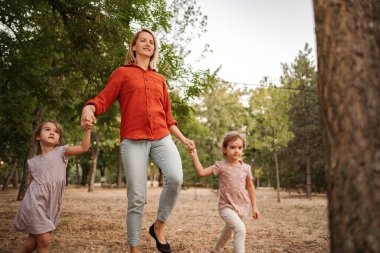 Anne, iki küçük kızıyla parkta yürümekten zevk alıyor, ellerini tutuyor.