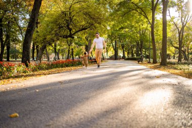 Güneşli bir yolda neşeli bir etkileşim başlar. Bir adam ve çocuk, canlı ağaçlar ve çiçek açan çiçeklerle çevrili bir parkta birlikte koşarken ve yanında mutlu bir köpek koşarken..
