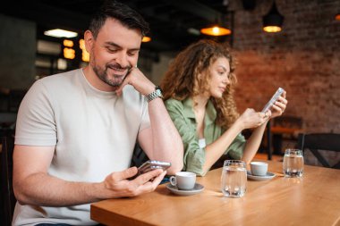 Kadın ve erkek, kahve dükkanındaki masada otururken akıllı telefonlarını kullanıyorlar.