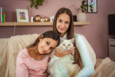 Two sisters and a fluffy cat bonding together on a couch at home, looking at camera