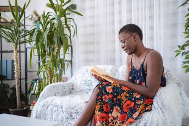 Young woman relaxing on a sofa reading a book in her cozy living room surrounded by plants