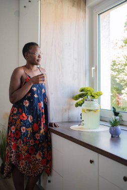 Serene young woman enjoying a glass of infused water while looking out the window in her modern kitchen
