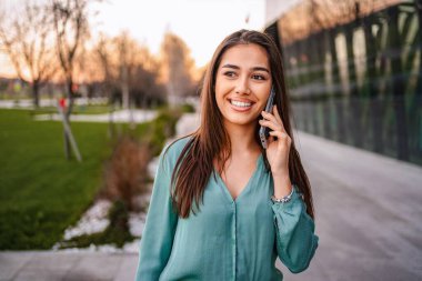 Cheerful businesswoman talking on smartphone while walking near office building