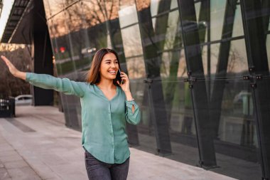 Smiling businesswoman hailing a taxi while talking on a smartphone in front of a modern glass building