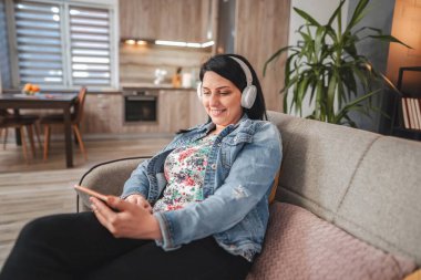 Mid adult woman relaxing on the sofa at home, listening to music with headphones and using a smartphone
