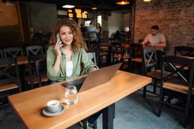 Bir kafede uzaktan çalışan, telefonla konuşan ve dizüstü bilgisayar kullanan genç bir kadın.