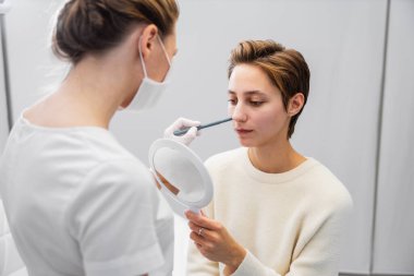 Makyaj sanatçısı, müşterisi aynadaki yansımasını incelerken temeli dikkatlice uygular..