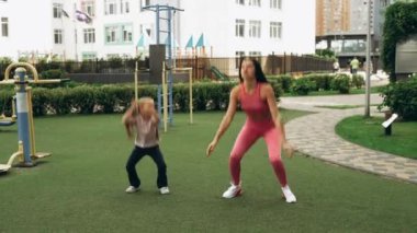 Bir anne ve kız birlikte dışarıda egzersiz yapıyor, oyun parkında çömelme hareketleri yapıyorlar..