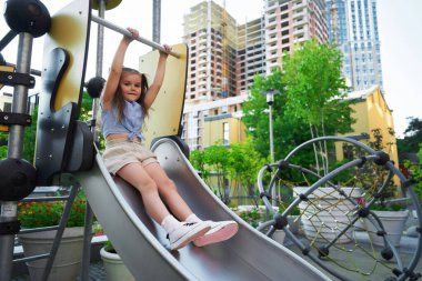 Bir kız şehir ortamında oyun parkında neşeli bir ifadeyle kayıyor..