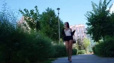 Bir kadın canlı bir park yolunda yürüyor, şık bir kıyafetle güneşli havanın tadını çıkarıyor..