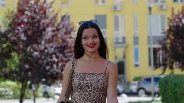 Gülümseyen bir kadın yeşil yapraklarla çevrili canlı sarı bir binanın önünden güvenle geçer..