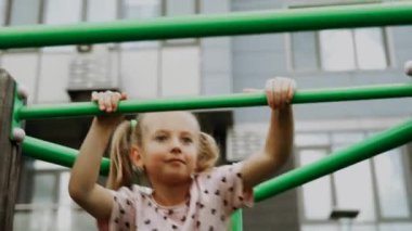 Bir kız, arka planda binalar olan oyun parkındaki parmaklıklara tırmanmaktan hoşlanır..