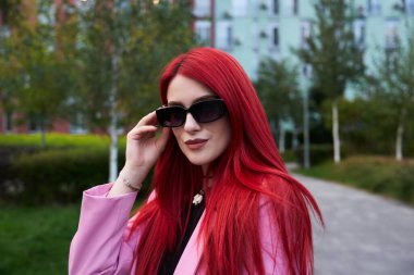 Pembe ceketli kadın güneş gözlüğünü güneşli bir parkta ayarlıyor..